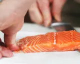 Hände halten ein Lachsfilet, während ein Messer die Haut vom Lachs entfernt.