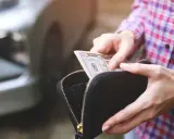 Ręce wyciągają banknot 50 dolarów z portfela. W tle widać fragment samochodu, sugerując kupno samochodu za gotówkę.