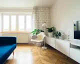 Jasny salon z niebieską kanapą i roślinami. Zasłony z wzorem pomogą jak wyciszyć pokój.