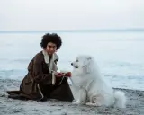 Kobieta z burzą włosów i biały pies siedzą na plaży. Spokojne morze w tle. Idealny dzień nad morze z psem.