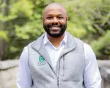 A smiling man with a beard wears a grey vest over a white shirt. The vest has a Tuck at Dartmouth logo, suggesting a curated professional image.