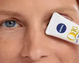Un tube de crème pour les yeux Nivea Q10 Anti-Wrinkle Power est tenu devant l'œil d'une femme, masquant les rides pattes d'oie.