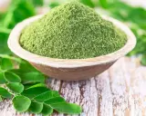 Moringa w proszku w drewnianej misce, otoczona świeżymi liśćmi. Zanim zaczniesz stosować, sprawdź moringa przeciwwskazania.
