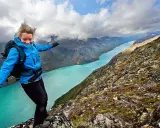 En kvinna med ryggsäck balanserar på en klippkant med en turkos sjö och berg i bakgrunden, en vy som påminner om Besseggen i Norge.