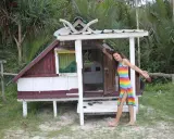 Kobieta w tęczowej sukience stoi obok małego, drewnianego domku na plaży.