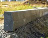 Gotowy do montażu betonowy krawężnik, idealny jako podbudowa pod obrzeża. Widać jego szarą, chropowatą powierzchnię i piasek pod nim.