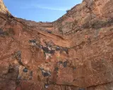 Acantilado de roca rojiza con vetas oscuras, mostrando diferentes tipos de canteras y la estratificación geológica bajo un cielo azul.