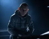 Młody mężczyzna w szarej bluzie z kapturem, z ogoloną głową, wygląda jak jeden z popularnych raperów, gotowy do występu.