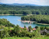 Malowniczy widok na jezioro z żaglówkami, otoczone lasami. W oddali widać góry, a na pierwszym planie domki letniskowe. Idealne miejsce na wypoczynek nad wodą, przypominające solina plaża.