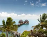 Dwa domki na wodzie połączone pomostem, otoczone palmami. Spokojne morze i błękitne niebo z chmurami. Zanzibar, pora deszczowa, ale pogoda dopisuje.
