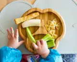 Babyhände greifen nach leckeren Baby-Snacks ab 6 Monaten: Banane, Gurke, Nudeln und ein Stück Brot auf einem Holztablett.