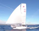 Un voilier de la classe mini, numéro 946, navigue sur une mer bleue sous un ciel clair.