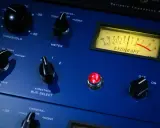 Gros plan sur un compresseur audio bleu avec des boutons, un VU-mètre et une lumière rouge allumée.