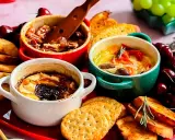 Drei kleine Auflaufförmchen mit geschmolzenem Camembert, garniert mit Nüssen, Tomaten und Kräutern, serviert mit Brot und Kirschen.