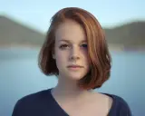 Ritratto fotografia di una giovane ragazza con capelli rossi e occhi azzurri, sullo sfondo di un lago e colline.