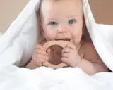 Ein Baby mit blauen Augen kaut auf einem Holzring. Vielleicht ist das der erster zahn baby, der durchbricht.