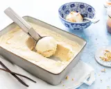 Une cuillère prélève une boule d'une glace vanille maison crémeuse dans un plat. Des gousses de vanille et des éclats de cacahuètes sont à proximité.