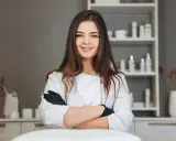 Uśmiechnięta młoda kobieta w białym fartuchu i czarnych rękawiczkach, gotowa do pracy w gabinecie. Marzysz o karierze w kosmetologii? Studia w Warszawie czekają!