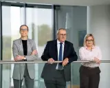 Trzy osoby w strojach biznesowych stoją przy balustradzie w nowoczesnym wnętrzu.