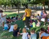 Niños sentados en el césped, participando en una gymkana para niños. Un adulto con camiseta amarilla dirige la actividad.