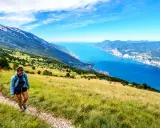 Zwei Wanderer genießen die Aussicht beim Monte Baldo Wandern. Der Weg führt über einen sonnigen Hang mit Blick auf den tiefblauen Gardasee.