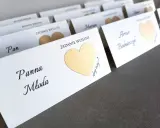 Białe karty z imionami gości i złotymi serduszkami kryją śmieszne zadania dla gości weselnych.