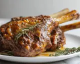 Trois souris d'agneau braisées, nappées d'une sauce dorée et garnies de romarin frais, sur une assiette blanche.