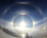 Zjawiskowy efekt halo nad zimowym krajobrazem. Słońce otoczone tęczowymi kręgami, mgła i ośnieżone drzewa tworzą magiczną scenę.