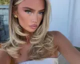 Kobieta z blond włosami, stylizowanymi na fale, z subtelnym makijażem i złotymi kolczykami. Jej fryzura przypomina buzz cut, ale jest dłuższa i bardziej objętościowa.