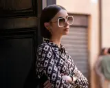 Emily in Paris, Lily Collins poses with large white sunglasses and a patterned shirt, leaning against a dark door.