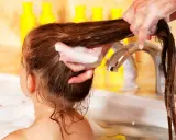 Schaumige Hände waschen lange, verfilzte Haare von Kindern in der Badewanne. Gelbe Flaschen im Hintergrund.