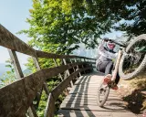 Ciclista in azione durante il giro del lago di Molveno in bici, su un ponte di legno tra alberi verdi e montagne.