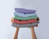 Manos sostienen una pila de ropa doblada en tonos pastel. Consejos para saber como evitar que una prenda destiña.