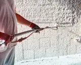 Pracownik w rękawicach wykonuje tynkowanie ścian za pomocą agregatu. Powstaje chropowata, biała powierzchnia.