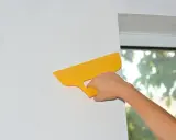 Ręka z żółtą szpachlą wygładza tapetę przy oknie. Dowiedz się, jak kłaść tapetę od okna czy do okna, by efekt był idealny.