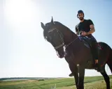 Uomo con casco e barba cavalca un cavallo scuro in un paesaggio verdeggiante. Un'esperienza di **Toscana a cavallo**.