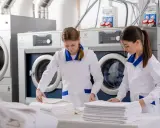 Pracownice hotelowej pralni składają czyste ręczniki. W tle widać profesjonalne pralki i suszarki.