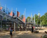 Un día perfecto de **munich con niños** en un parque infantil moderno con toboganes y estructuras de juego, con torres de iglesias al fondo.