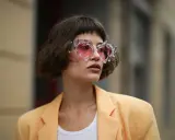 Frau mit gestuftem Bob mit Pony und auffälliger Brille mit rosa Gläsern, trägt einen hellgelben Blazer.