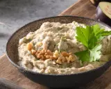 En skål med krämig baba ganoush, toppad med valnötter och persilja. Bredvid ligger skivade auberginer och ett vitlöksklyfta.