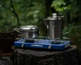 Niebieska kuchenka turystyczna Campingaz z dwoma palnikami, na której stoją metalowy garnek i kawiarka, gotowa do przygotowania posiłku w lesie.