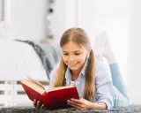 Uśmiechnięta dziewczynka leży na dywanie i czyta książkę. Może odkrywa w niej mądrość przysłowia.