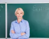 Nauczycielka angielskiego z uśmiechem, stojąca przed tablicą z napisem "English". Marzy o wcześniejszej emeryturze dla nauczycieli.