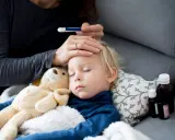 Bambino addormentato con febbre, la mamma controlla la temperatura. Vicino ci sono flaconi di antibiotico.