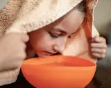 Kind inhaliert Dampf aus einer Schüssel, um bei Bronchitis Linderung zu finden. Ein Handtuch bedeckt den Kopf.