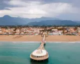 Un molo si estende su un mare azzurro, con una spiaggia dorata e una città costiera sullo sfondo. La Toscana offre un mare bello e panorami indimenticabili.