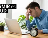 Joven con dolor de cabeza frente a su laptop, buscando trucos para dormir mejor. El reloj marca la hora.