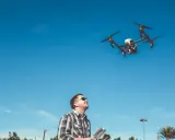 Ein **Drohnenpilot** steuert mit einer Fernbedienung einen Quadrocopter am blauen Himmel.