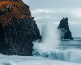 Vågor slår mot en klippvägg med basaltpelare och orange vegetation. Framför syns en mörk, svart sandstrand.