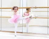 Mała baletnica w różowej tutu ćwiczy przy drążku. Jej odbicie w lustrze pokazuje, jak pilnie uczęszcza na lekcje baletu.